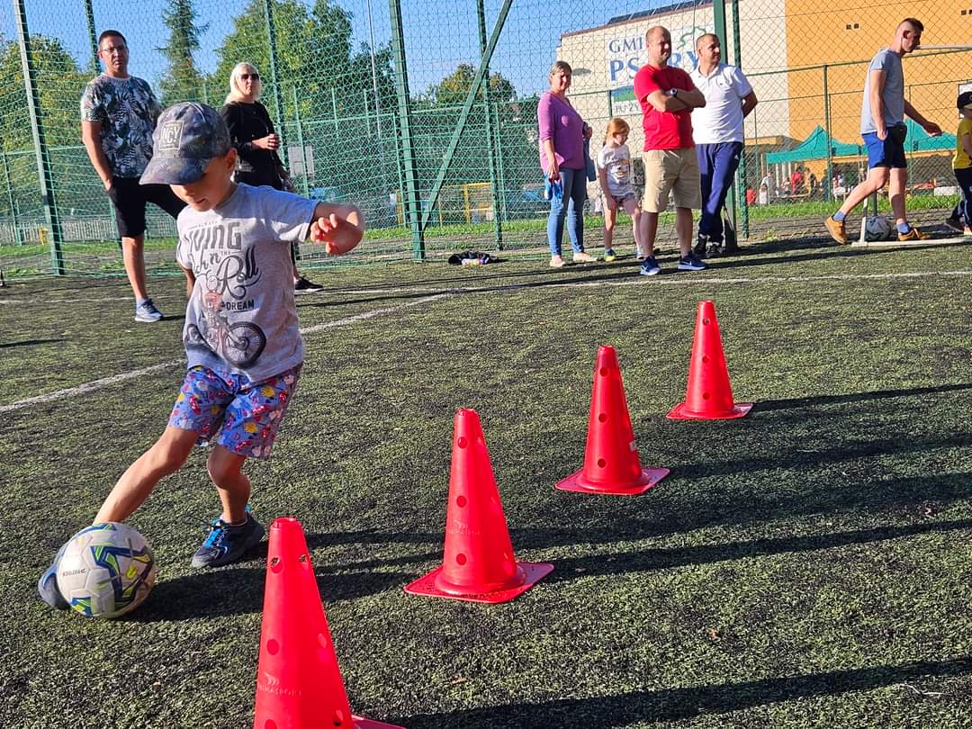 Na zdjęciu znjaduje się chłopczyk, który wykonuje ćwiczenie sprawnościowe z piłką nożną. Kieruje piłkę między czerwonymi pachołkami.